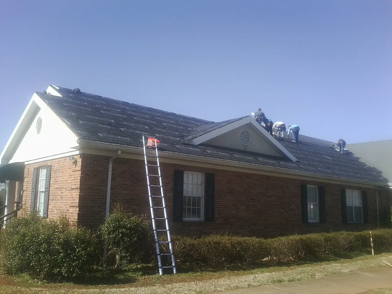 Crew tearing shingles off a brick ranch roof with a ladder set — Metal Roof Repair work by our crew in Edelstein
