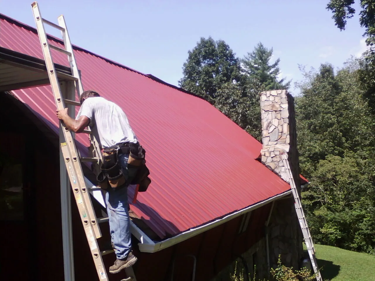 Roofer on a ladder beside a red metal roof and stone chimney — Soffit & Fascia Repair by Roofing Brothers in Edelstein